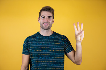 Young handsome man wearing casual blue t-shirt over yellow isolated yellow background showing and pointing up with fingers number three while smiling confident and happy tree