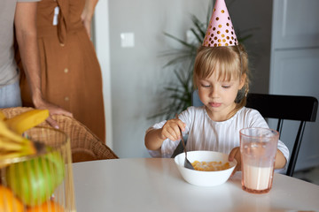 pretty adorable girl sitting in the kitchen alone, in party hat, before birthday party at home. eat, have meal in the morning