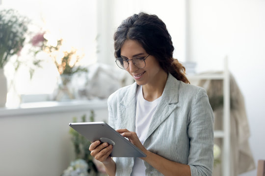 Head shot smiling young beautiful businesswoman decorator clothing designer in glasses using digital computer tablet, web surfing information or consulting clients online in modern creative office.