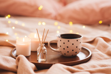 Cup of coffee with home aroma stickes and burning candles in bed closeup over glowing christmas lights. Winter holiday season.