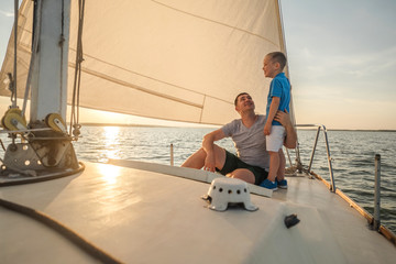 Happy traveler father and son enjoying sunset from deck of sailing boat moving in sea at evening...