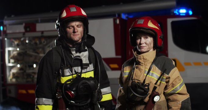 Middle plan of Caucasian man and woman firefighters in the full equipped costumes and helmets standing outside looking into camera at night. Concept of saving lives, heroic profession, fire safety
