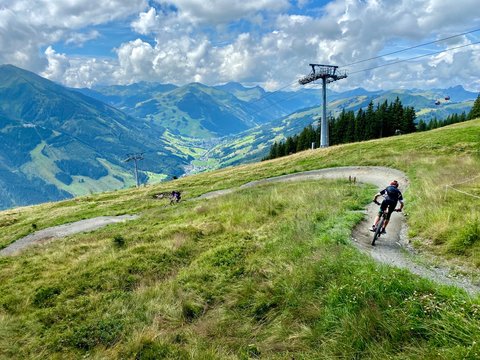 Unrecognicable bikers on a mountainbike trail in Saalbach in the Austrian Alps in summer with dramatic sky and cable car in the background.