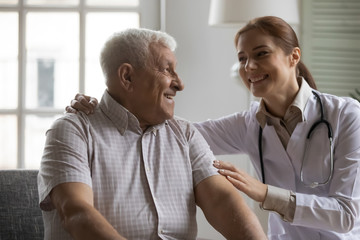 Obraz premium Smiling kind young nurse cuddling shoulders of elderly mature retired male patient, supporting indoors. Happy old 80s man enjoying conversation with pleasant doctor, medical insurance concept.
