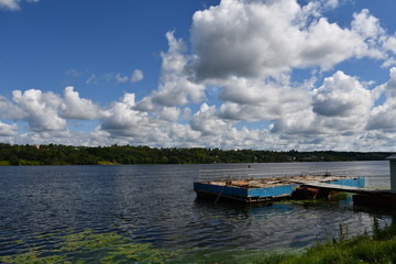 Fototapeta premium Panorama of a large river or lake. Water lilies grow near the shore. A small old dilapidated pier for boats and motorboats. Beautiful clouds.