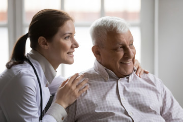 Dreamy young female doctor cuddling shoulders of smiling old retired patient, looking together at distance. Head shot happy caring nurse supporting elderly mature man, thinking of healthy lifestyle.