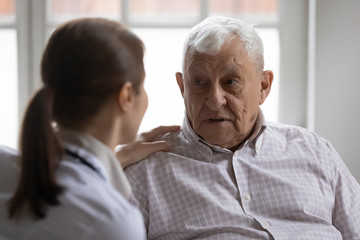 Head shot sad older 80s man sharing health problems with young nurse at home visit. Female doctor gp giving professional psychological help to unhappy elderly retired patient, medical service concept.