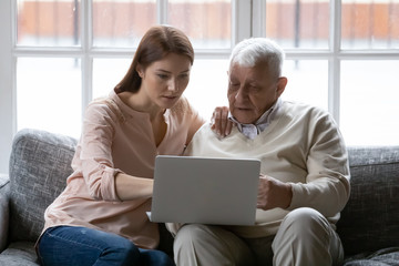 Focused young woman helping older daddy with computer applications, sitting together on comfortable sofa in living room. Serious different generations enjoying web surfing information in internet.