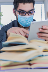 A concentrated student, wearing a protective mask, reads from a tablet and flips through the pages of a textbook, along with other open books on the desk, studying from home. Long distance education.