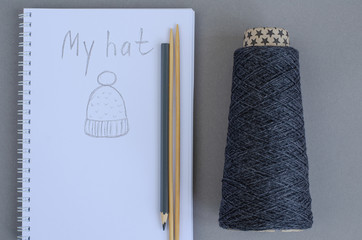 dark gray yarn cone, bamboo knitting needles, pencil and notebook with sketch hat. gray background. Knitting hobby craft concept.