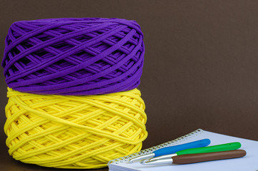 yellow and purple t-shirt yarn balls and colorfull crochet hooks on notebook on brown background. Craft, handmade, hobby concept.