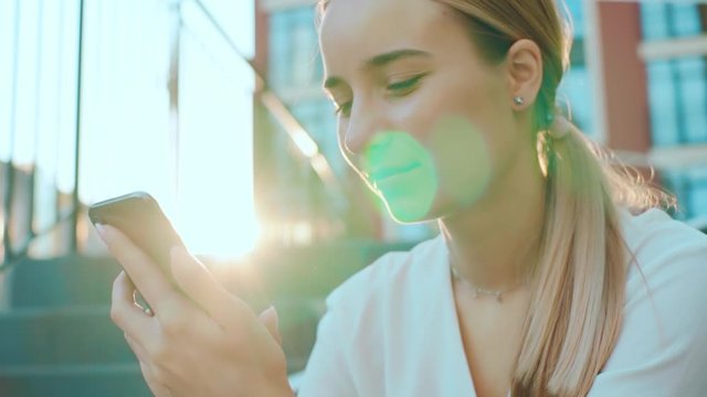 At sunlight young woman sitting use phone smiling feel happy on street outdoors girl message female outdoor smartphone street attractive city portrait internet slow motion