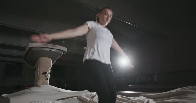 Girl gymnast doing a straight body flip or somersault off a raised platform landing on mats in a dark gym with single bright spotlight. Night gymnastics training of a girl in a sports hall at night.
