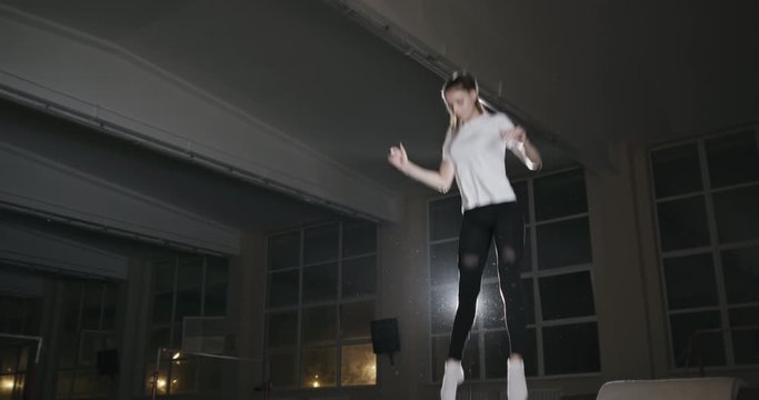 Fit athletic young woman bouncing high on a trampoline backlit by a bright spotlight in a gym. Night gymnastics training of a girl in a sports hall at night.