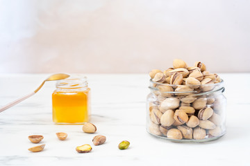 A transparent jar of pistachios stands on a white table. Nearby is a small jar of honey and a few peeled nuts and shells. Vegetarian food. Raw food concept. Healthy dessert