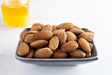 Peeled almonds in a rectangular gray bowl stand on a white table. There is a jar of honey nearby. Healthy food, tasty and healthy snacks. Foods are high in antioxidants, minerals and vitamins. 