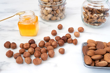 Peeled almonds are scattered on a white table. There are jars of honey, walnuts, pistachios and a bowl of almonds nearby. Healthy food high in antioxidants, protein and vitamins