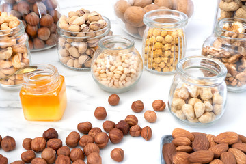 An assortment of different nuts in glass jars: hazelnuts, cashews, pistachios, pine nuts, walnuts, almonds and chickpeas. Jars of nuts are on a white table, near a small jar of honey