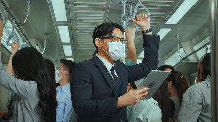 Asian businessman wear mask while traveling to work on skytrain. Prevent Coronavirus, bacteria in public place from people. Business people use tablet outside meeting at peak hour no social distancing