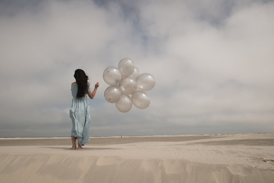Young woman in blue dress with white balloons on beach on windy day
