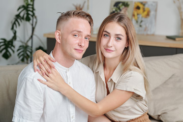 Young blond pretty woman embracing her husband while both sitting on sofa