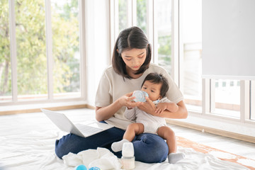 Close up of Young asian mother on maternity leave trying to freelance by the desk with toddler child.Stay at home mom working remotely on laptop while taking care of her baby.