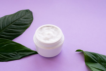Soft white cream in jar with bright green leaves on purple background. Spa treatment and procedure at home.
