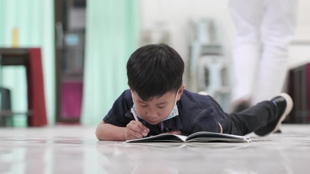 Asian boy about 4 years old with face mask under his jaw drawing a picture by pencil while lying on the floor