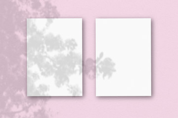 2 vertical sheets of textured white paper on pink table background. Mockup overlay with the plant shadows. Natural light casts shadows from an exotic plant. Horizontal orientation