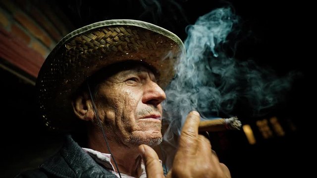 The man smokes a cigar. An elderly farmer in a straw hat smokes a cigar outside the ranch. Confident Senor reflects in cigarette smoke. An old cowboy in a leather vest. Evening rest near the house.