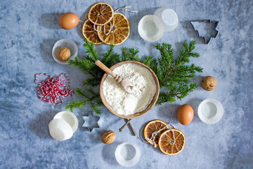 Culinary Christmas food background. Baking ingredients, flour, eggs, cookie cutters, fir branches, dried oranges, nuts, paper muffin molds on a dark table. Top view, flat lay