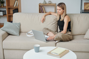 Contemporary young blond woman with crossed legs using laptop at home