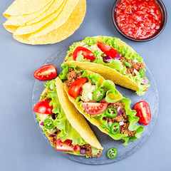 Taco shells with lettuce, ground beef meat,  mashed avocado, tomato, red onion and jalapeno pepper, on stone plate, square format, top view