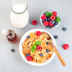 Small cereal pancakes with maple syrup, raspberry, blueberry and milk, in  white bowl, horizontal, top view, square