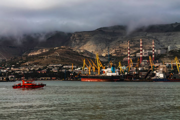 A red harbor tug sailing in the bay against the background of a cargo ship moored in the seaport against the background of mountains and factory pipes. Cloudy weather in the mountains.