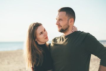 Beautiful woman full of love looking to handsome convinced man embracing her. Young adult couple standing on coast and huging each other. Millennial male and female posing on beach on sunny day