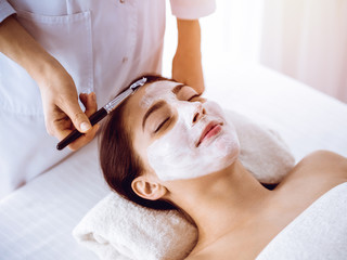 Beautiful brunette woman enjoying applying cosmetic mask with closed eyes in sunny spa center. Relaxing treatment and medicine concept