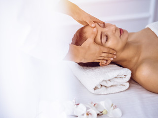 Fototapeta na wymiar Beautiful brunette woman enjoying facial massage with closed eyes in sunny spa center . Relaxing treatment and cosmetic medicine concepts