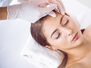 Fototapeta na wymiar Beautician doing beauty procedure with syringe to face of young brunette woman in sunny spa center. Cosmetic medicine and surgery, beauty injections