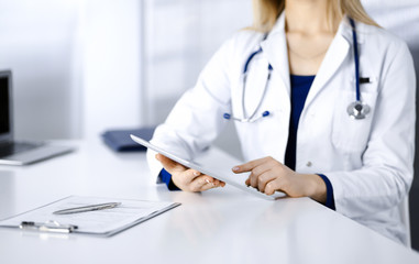 Unknown young woman-doctor is checking some medication names, while sitting at the desk in her cabinet in a clinic. Physician with a stethoscope is using a tablet computer, close-up. Perfect medical