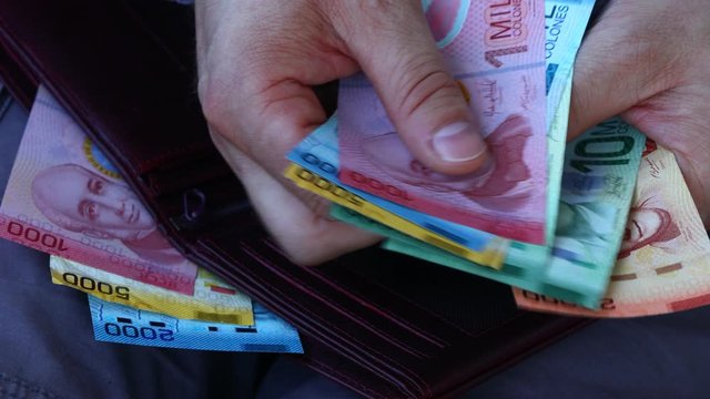 A man in a business shirt counts Costa Rica money, Colones, Various banknotes. The stuffed wallet in his lap