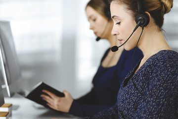 Two busineswomen have conversations with the clients by headsets, while sitting at the desk in a modern office. Diverse people group in a call center. Telemarketing and customer service