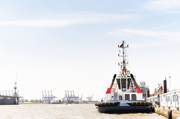 Naklejka premium HARBOR OF HAMBURG, GERMANY, May 31, 2020: Tug ship VB Resolut moored to the quay wall with industrial port and cranes in the background.