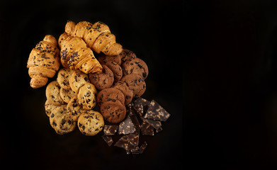 Tasty golden croissants on black background. Freshly baked chocolate chip cookies with broken milk chocolate pieces. Closeup of a group of French pastry from bakery .Top view with Copy space. 