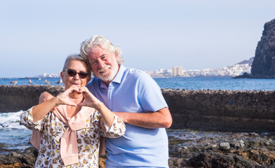 Senior couple with white hairs hugging at the beach making heart shape with hands - horizon over water and city - concept of active seniors during holidays