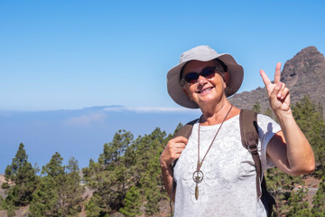 Happy senior woman with hat and backpack enjoying excursion in mountain range in Tenerife island making ok sign- concept of vacation into the nature for an elderly active people