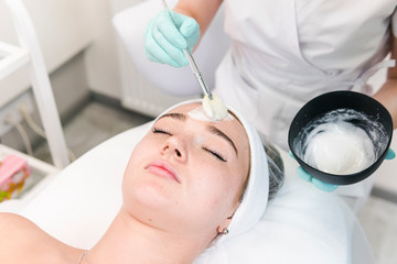 Naklejka premium The young female client of cosmetic salon having a cleansing facial mask. The procedure of applying a peeling mask to the face. Concepts of skin care and beauty salon or clinic.