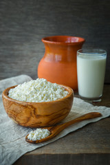 Fresh home-made curd in a wooden bowl on a linen napkin, with a glass of milk and a clay jug on a wooden old background.