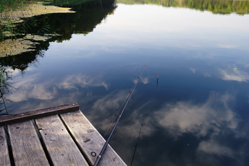 fishing rod on a wooden pier and reflection of clouds in the lake water