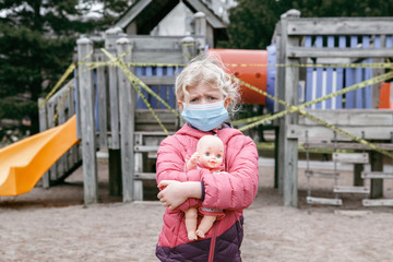 Sad Caucasian girl in face mask with baby toy on closed playground outdoor. Kids play area locked with yellow caution tape in Toronto city, Canada. Coronavirus social distance quarantine. - 373324549